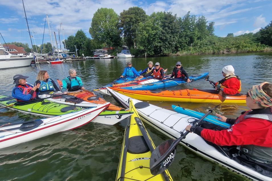 Gruppe von Kajakfahrern in bunten Kajaks versammelt sich auf Gewässer nahe einem Yachthafen unter blauem Himmel.
