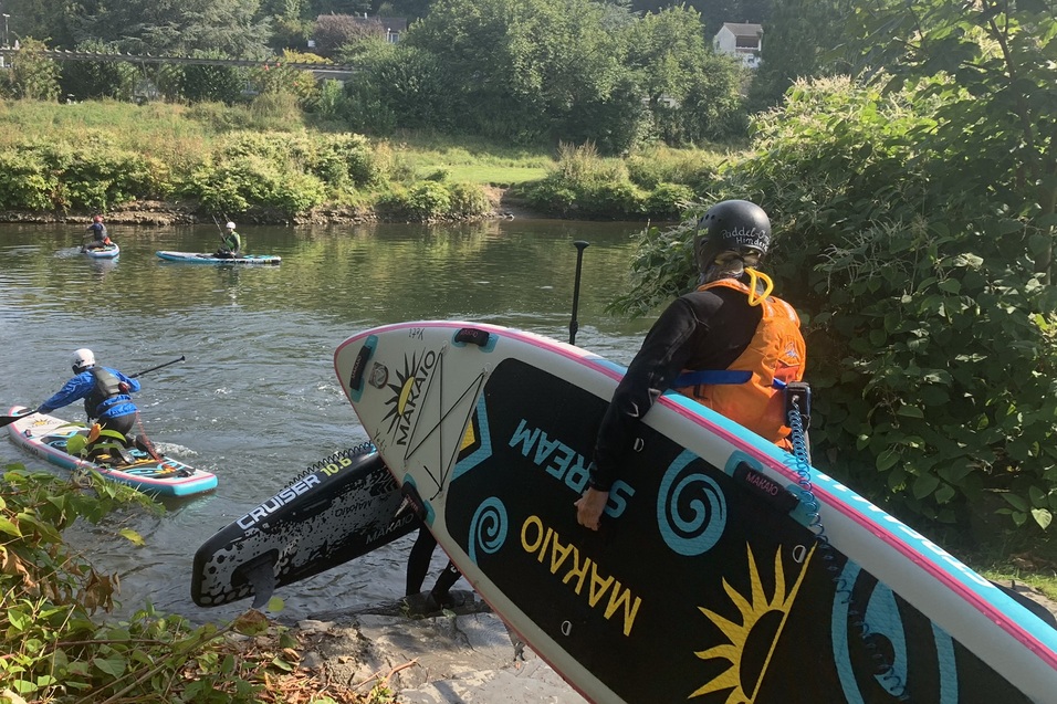 Person trägt ein Stand-Up-Paddle-Board zu einem Fluss, während andere Paddler im Wasser unterwegs sind.