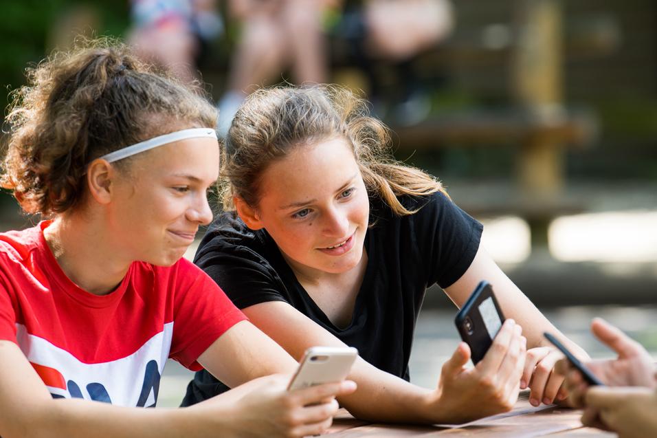 Zwei Mädchen sitzen an einem Tisch, schauen auf ihre Smartphones und lächeln dabei. Im Hintergrund sind weitere Personen sichtbar.