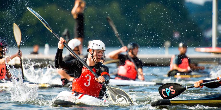 Wettkampf im Kanusport mit mehreren Athleten in roten Westen, paddelnd auf einem Gewässer. Spritzendes Wasser ist sichtbar.